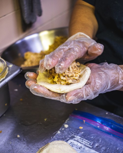 A person in gloves holds a dough piece filled with shredded chicken, preparing it for cooking in a kitchen setting.
