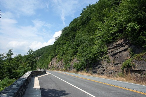 A winding road alongside a rocky cliff, surrounded by lush green trees under a blue sky.