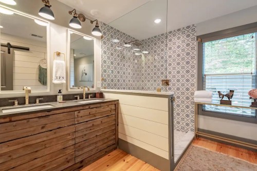 Modern bathroom featuring a glass shower, patterned tile wall, wooden vanity, and natural light from a window.