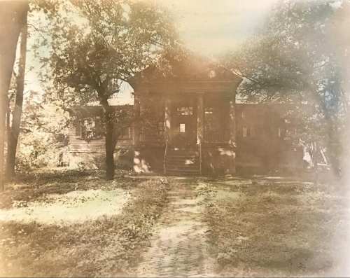 A vintage photo of a large, old house surrounded by trees, with a pathway leading to the entrance.