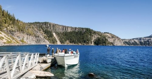 A boat docked at a scenic lake with people boarding, surrounded by lush green hills and clear blue water.
