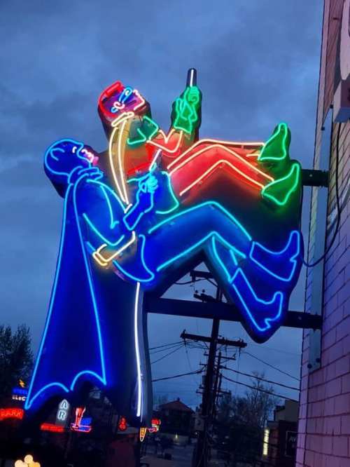 Neon sign featuring two cartoonish figures, one in a red outfit and the other in a blue cape, sitting together.