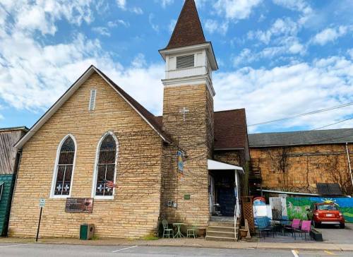 A stone church with a tall steeple, surrounded by a blue sky and clouds, featuring outdoor seating in front.