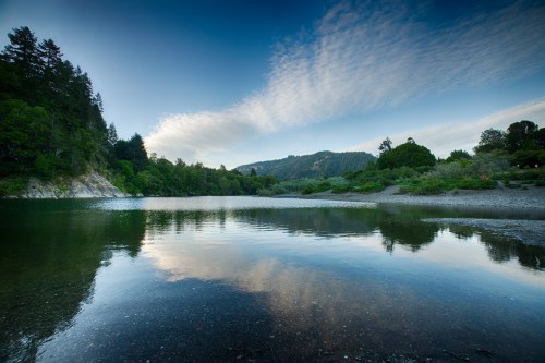 A serene river scene at dusk, reflecting trees and clouds under a blue sky, surrounded by lush greenery.