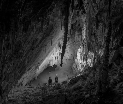 Two explorers with headlamps stand in a dark cave, surrounded by stalactites and stalagmites, illuminated by soft light.