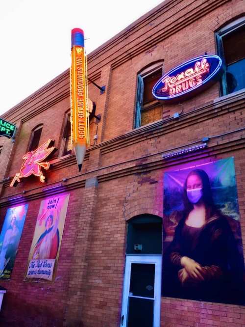 A colorful building facade featuring neon signs, a large pencil, and a masked Mona Lisa mural.