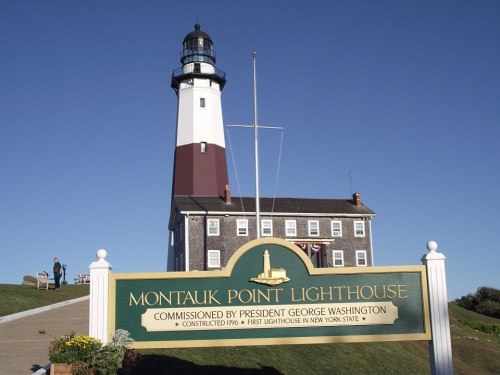 Montauk Point Lighthouse with a sign detailing its history, set against a clear blue sky.
