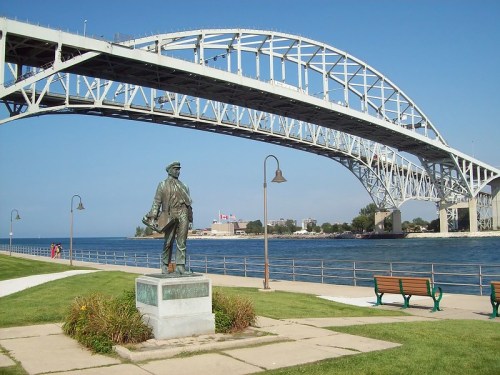 A statue of a man stands by the water, with a large bridge arching overhead and a clear blue sky in the background.
