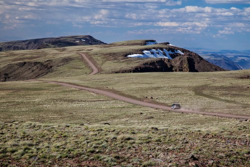 A dirt road winds through a grassy landscape with distant mountains and patches of snow under a blue sky.