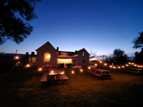 A cozy outdoor setting at dusk, featuring picnic tables and string lights illuminating a charming house.