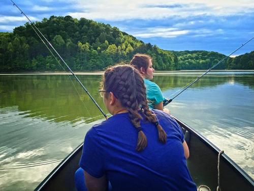 Two people fishing from a boat on a calm lake, surrounded by lush green hills and a cloudy sky.
