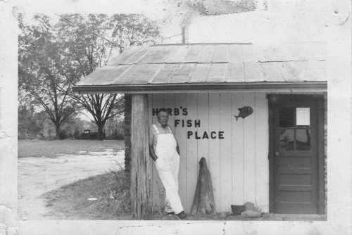 A man in overalls stands beside a rustic building with a sign reading "Herb's Fish Place" in a rural setting.
