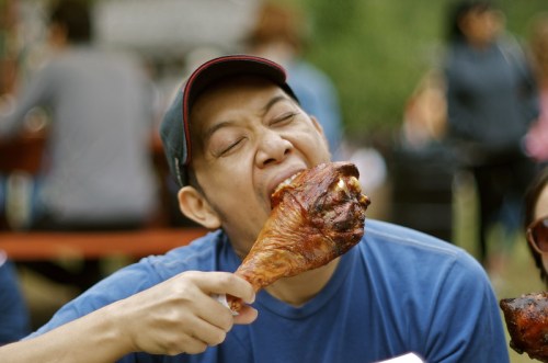 A man in a blue shirt and cap joyfully bites into a large roasted turkey leg outdoors.