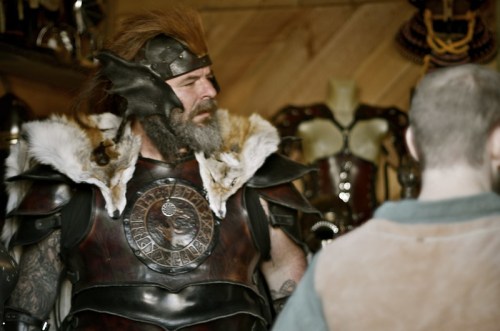A man in elaborate armor with a fur collar and a beard stands in a shop filled with medieval-style attire and accessories.