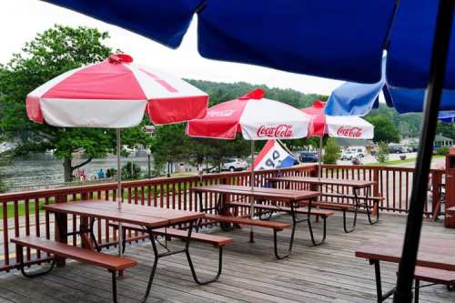 Outdoor seating area with wooden picnic tables and red and white umbrellas by a lake.