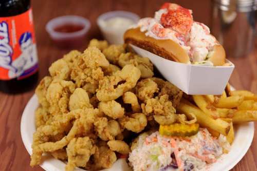 A plate of fried seafood, including crispy fried shrimp, a lobster roll, fries, coleslaw, and a soda.