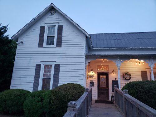 A charming white house with a porch, surrounded by greenery, illuminated by warm lights at dusk.