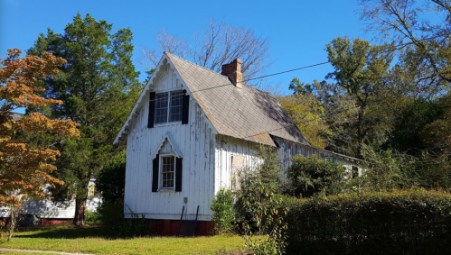 A weathered white house with a steep roof, surrounded by trees and shrubs under a clear blue sky.