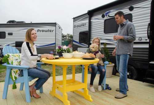 A group of three people enjoying a meal outdoors by a camper, with a dog and a bright yellow table.