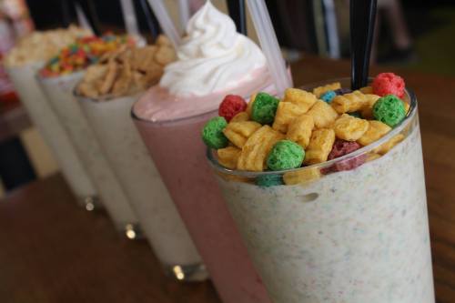 Colorful milkshakes topped with cereal and whipped cream, lined up on a wooden table.