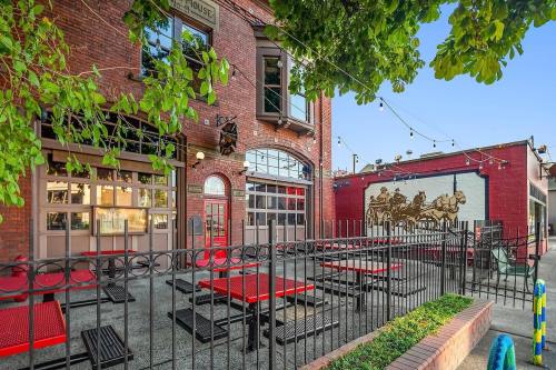 Outdoor seating area of a brick building with a mural, surrounded by greenery and string lights.