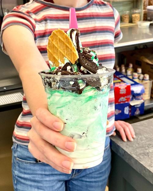 A person holds a colorful milkshake topped with whipped cream, chocolate syrup, a cookie, and a waffle cone.