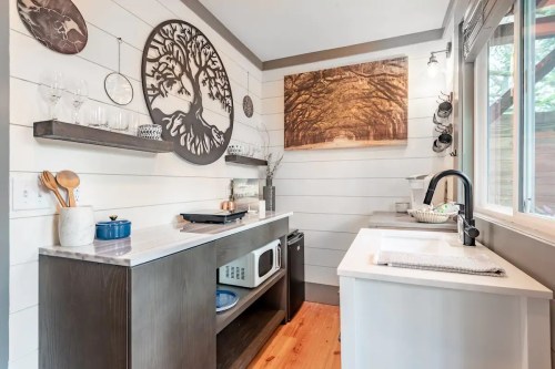 A modern kitchen with white shiplap walls, wooden accents, and a tree-themed wall art, featuring a sink and microwave.