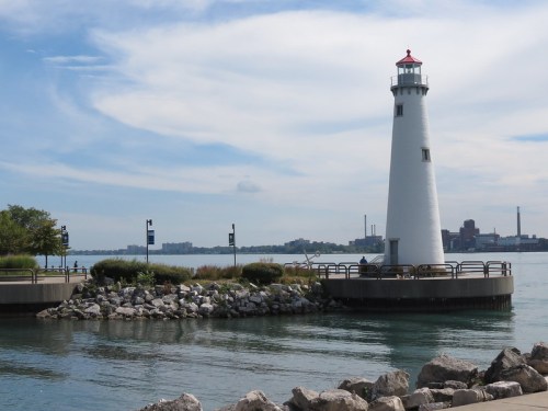 A white lighthouse stands by a calm waterway, surrounded by rocks and greenery under a blue sky with clouds.