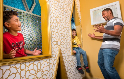 A man joyfully interacts with two children in a colorful, playful room with unique mirrors and patterns.