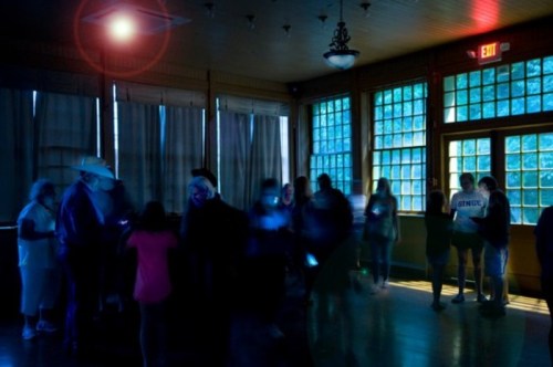 A dimly lit room with people gathered, some using phones, illuminated by blue and red lights.