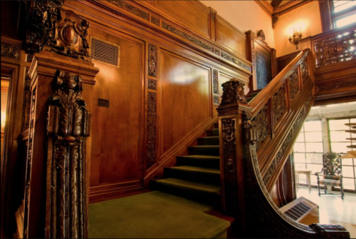 Elegant wooden staircase with intricate carvings and green carpet, set in a richly decorated interior.