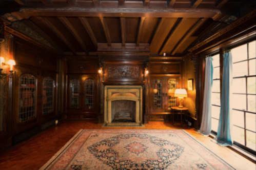 A cozy, wooden-paneled room with a fireplace, large windows, and an ornate rug on the floor.