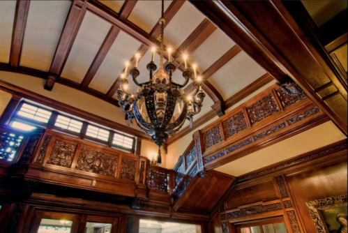 A grand interior featuring ornate wooden paneling, a detailed chandelier, and a staircase with intricate carvings.