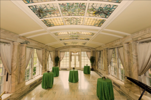 Elegant room with stained glass ceiling, marble floors, and green tablecloths on round tables. Natural light floods in.