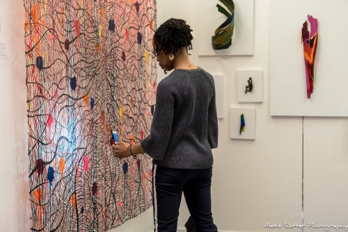 A person with short curly hair examines colorful artwork on a gallery wall, holding a phone to capture the piece.