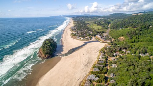 Aerial view of a coastal beach with waves, a rocky outcrop, and a nearby town surrounded by lush greenery.