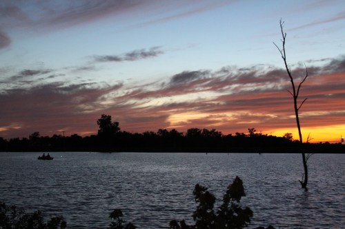 A serene sunset over a lake, with colorful clouds and silhouettes of trees, and a small boat in the distance.
