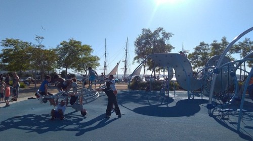 Children play on a colorful playground with sailboats in the background under a clear blue sky.