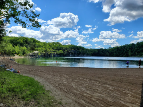 A serene lakeside scene with sandy shore, lush greenery, and fluffy clouds reflecting in the water.