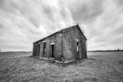 An abandoned brick building covered in vines, set against a cloudy sky and open field.