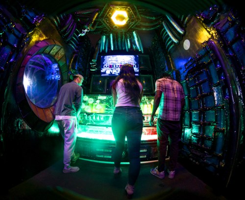 Three people interact with a colorful, futuristic arcade machine in a vibrant, sci-fi themed room.