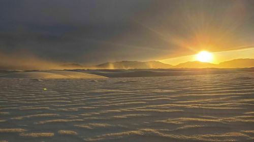 A serene desert landscape at sunset, with rippled sand dunes and soft light illuminating distant mountains.