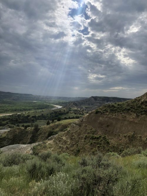A scenic view of rolling hills and a river under a cloudy sky, with sunlight breaking through the clouds.