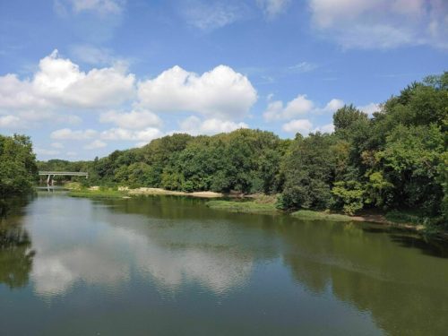 A serene river scene with lush green trees and a blue sky with fluffy clouds reflected in the water.
