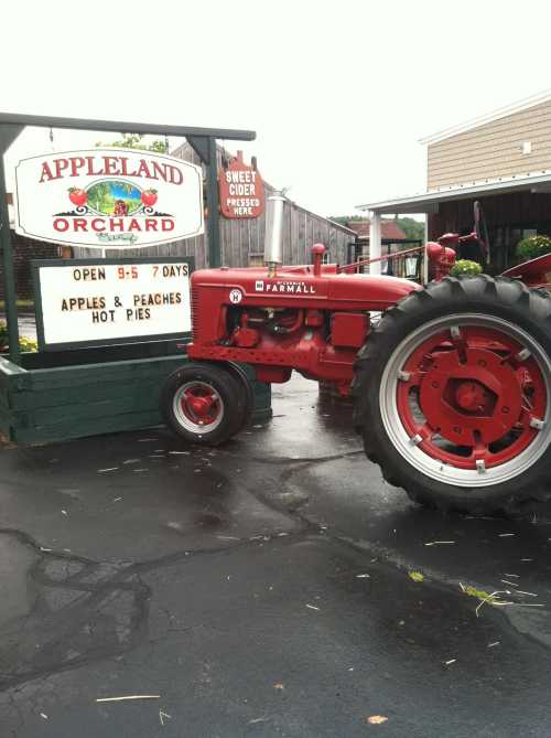 A red tractor parked beside a sign for Appletown Orchard, advertising apples, peaches, and hot pies.