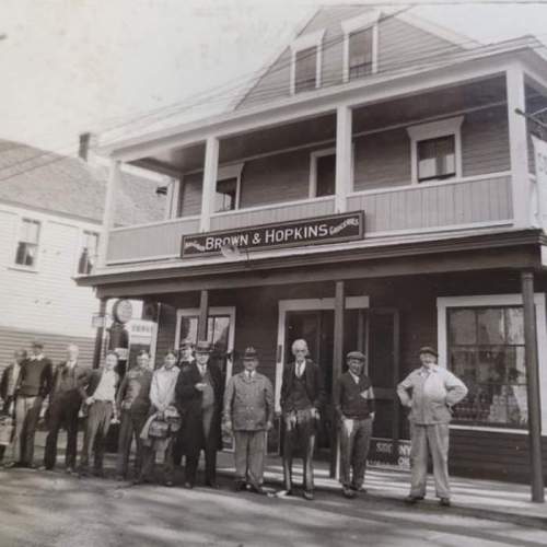 A historic black-and-white photo of a group of people standing outside a two-story building labeled "Brown & Hopkins."