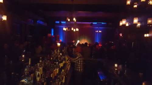 A dimly lit bar scene with a long counter, shelves of bottles, and a stage in the background with performers.