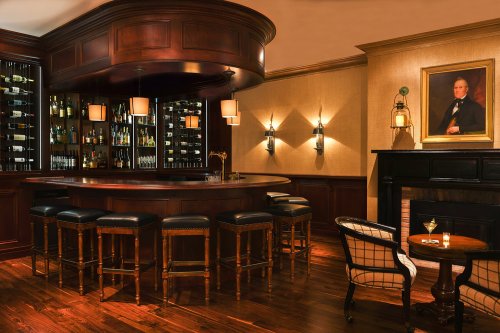 A cozy bar with a wooden counter, high stools, shelves of liquor, and a portrait on the wall, illuminated by soft lighting.
