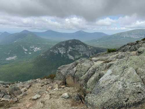 A rocky mountain summit with a panoramic view of green valleys and distant peaks under a cloudy sky.