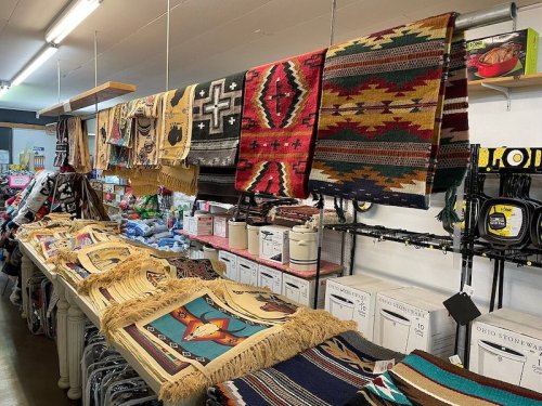 Colorful woven rugs and blankets displayed on shelves in a store, with various patterns and textures visible.